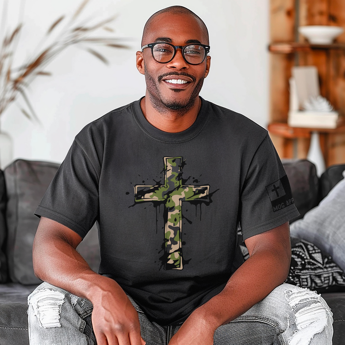 A smiling man wearing a pepper crewneck t-shirt with a green, black and cream camouflage cross graphic on the front, sitting on a couch in a living room setting.